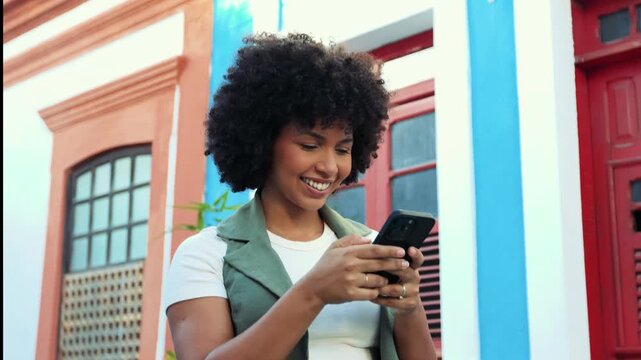 Young Black woman smiling while texting on her smartphone in a colorful colonial street. A moment of digital connection, joy and modern lifestyle.