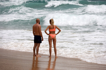 Tanned couple standing on a tropical sea beach on breaking waves background, man and girl in swimwear