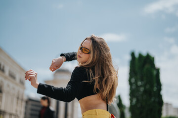 A fashionable woman wearing sunglasses dances energetically in an urban outdoor setting, expressing...