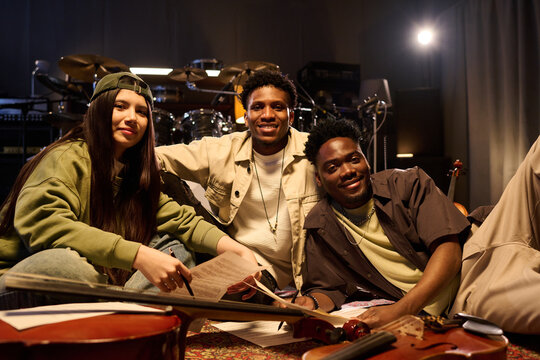 Group of young adult multiethnic musicians sitting on floor in music studio smiling at camera, holding musical instruments and sheet music, showing friendly teamwork and creativity