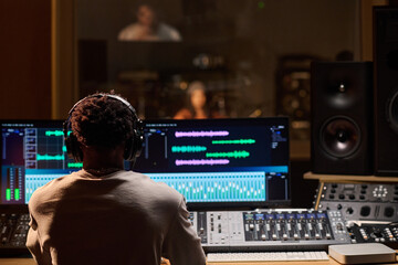 Black man wearing headphones operating audio mixing console and computer monitors in professional...