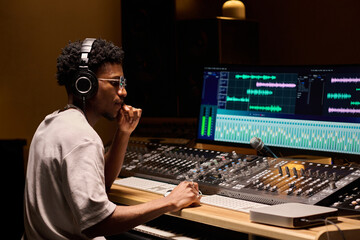 Young adult Black man wearing headphones working at audio mixing console in professional recording...