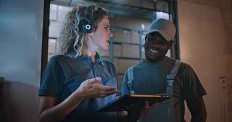 African American Worker and Female Manager Talking, Using Tablet Computer, Standing Outside of Logistics Retail Warehouse Full of Online Orders, Goods. Working in Express Delivery Service at Night.
