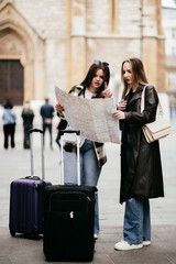 Two female tourists standing with luggage while reading a map and planning their route in the city.