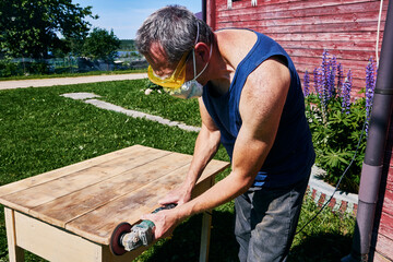 Working with handheld grinder on wooden tabletop, carpenter using protective gear during outdoor...
