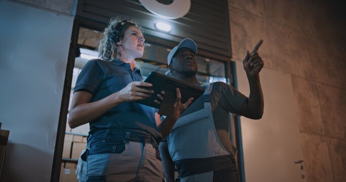 African American Employee and Female Manager Working in Express Delivery Service at Night: They Talking, Using Digital Tablet while Standing Outside of Logistics Warehouse Full of E-Commerce Goods. - Powered by Adobe