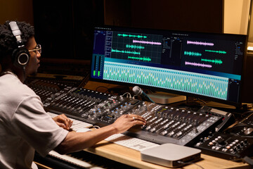Young adult Black man wearing headphones working at audio mixing console editing music tracks on...