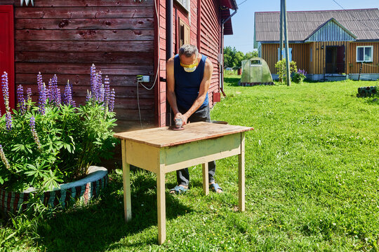 Using angle grinder for sanding wooden table, homeowner working on backyard project surrounded by grass, flowers and rural houses.