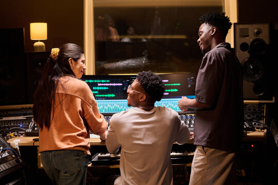 Multiethnic group of young adults collaborating in music studio, Black man operating audio mixing console while Hispanic woman and Black man standing nearby discussing project