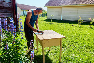 Sanding wooden tabletop with angle grinder outdoors, hobbyist working in yard near wooden houses on...