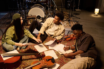 Multiethnic group of musicians collaborating on music composition, sitting on floor with violins...