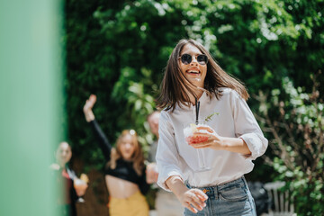 A cheerful young woman with sunglasses and a drink, surrounded by friends enjoying an outdoor party...