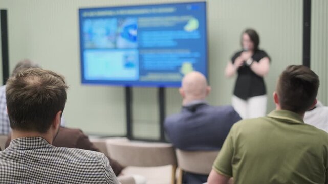 Audience members listen intently as a speaker presents information on a digital screen during a professional meeting or seminar in a corporate setting.