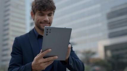 Portrait of happy Hispanic manager scrolls internet on digital tablet in urban environment. Young businessman reads online reports on device screen standing against skyscraper