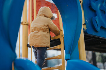 Young child wearing winter coat and pants climbs a bright playground ladder, showcasing energy and...