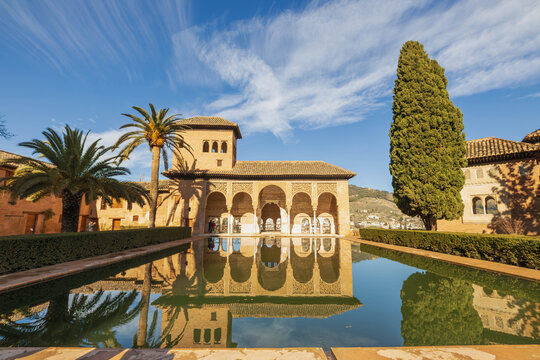 Frontal view of El Partal in the Alhambra, Granada, with its reflection mirrored in the water pool under a clear blue sky with few clouds.