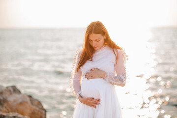 Happy pregnant woman 20-24 year old wearing white vintage dress standing over sea and sunset...