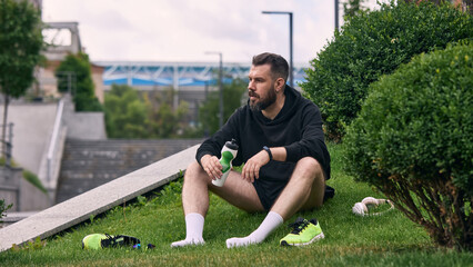 Man relaxing post-workout with water bottle on grassy urban hillside. Concept of active recovery, rest phase, urban athlete pause, hydration reminder, and training cycle balance.