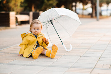 Smiling cute baby girl 3-4 year old wearing yellow raincoats and rubber boots with umbrella sitting on road in park outdoors. Autumn season. Childhood.