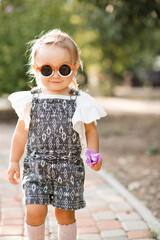 Funny cute baby girl 2-3 year old wearing sun glasses and stylish summer clothes in park outdoors. Looking at camera. Childhood.