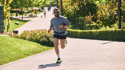 Man in headphones and sunglasses jogging through sunny park. Concept of confident pace, urban...