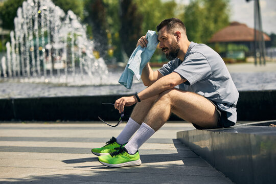 Man sitting by fountain taking break from exercise with towel and sunglasses. Concept of cooldown break, outdoor bench rest, relaxed runner, post-workout recovery, and athletic calm.