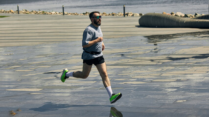 Middle-aged jogger runs along sunny waterfront in fitness attire. Concept of steady motion, urban...