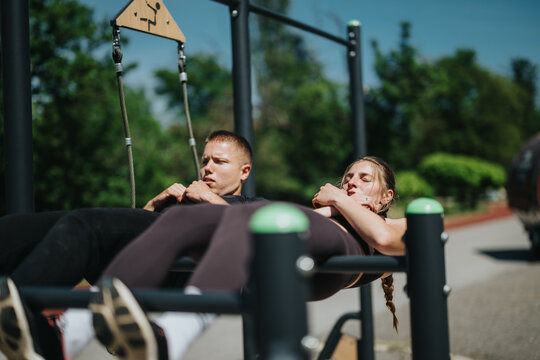 A man and a woman engage in core strengthening exercises at an outdoor fitness station surrounded by greenery. - Powered by Adobe