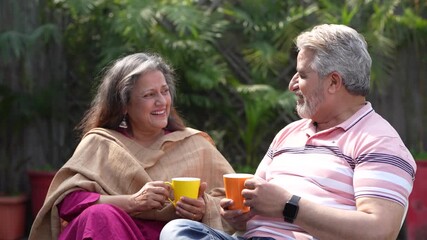 Happy Indian senior couple drinking tea or coffee in colorful mugs sitting at garden. - Powered by Adobe