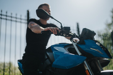 A person wearing black attire is leaning on a blue sports motorcycle on a sunny day. The background...