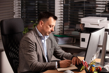 Confident businessman in a suit jacket working at a modern office desk. Sitting with a notebook and...