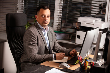 Confident businessman in a suit jacket working at a modern office desk. Sitting with a notebook and...