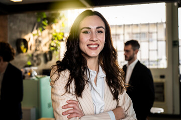 Portrait of a confident young businesswoman smiling with arms crossed in a modern office. She radiates leadership, ambition, and authenticity. Millennial professional and female empowerment concept.