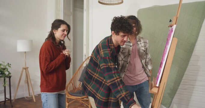 Three friends chat and try to find meaning in an abstract painting at home. Two guys and a girl are standing near a painting and canvas talking and having fun