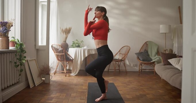 Close up of athletic girl in orange sweater doing yoga and meditation and standing in eagle pose on rug at home. The girl practices Garudasana in yoga