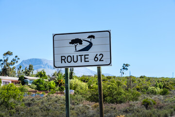 Ein weißes Schild mit dem Logo und der Aufschrift ROUTE 62 steht vor einer Landschaft mit Häusern, Bäumen und Bergen