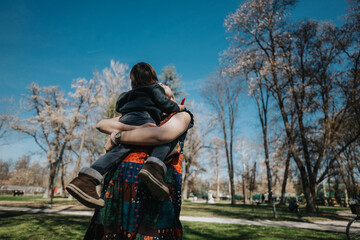 A woman holding a small child in an outdoor park. The trees are blooming in the background,...