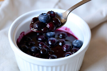 Spoon lifting homemade blueberry compote from a white ramekin, a delicious and sweet berry sauce perfect as a dessert topping for pancakes, yogurt, or ice cream