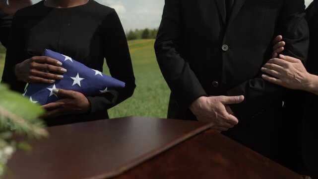 Midsection of unrecognizable family members or friends in black clothes standing near coffin at outdoor funeral holding folded American flag and grieving loss of loved one