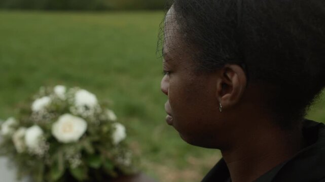 Side view close up shot of young African American widow wearing mourning veil wiping away tears from her face grieving loss of beloved husband at outdoor funeral