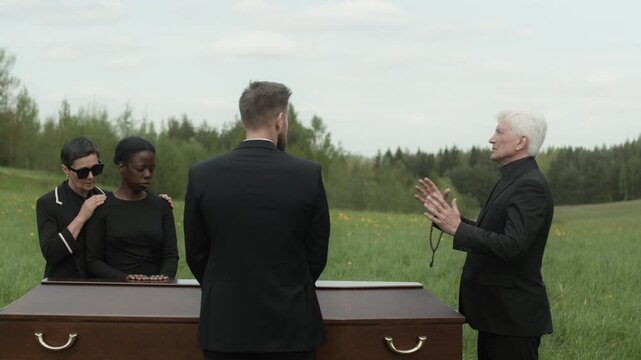 Medium long shot of senior Caucasian priest in black soutane holding rosary beads praying in front of closed casket at outdoor funeral ceremony with grieving multiethnic people
