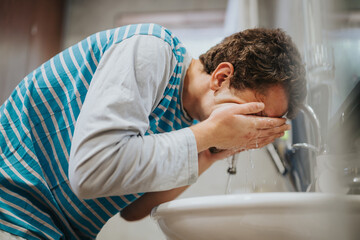 A young man in casual clothing washing his face over a bathroom sink, illustrating a refreshing...