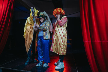 Three performers dressed in elaborate costumes interact on a stage under dramatic lighting and...