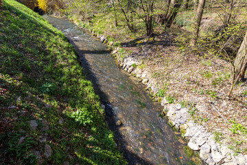 Gulp River with shallow flowing water seen from top perspective, stones on bank, green grass and trees with sparse foliage, sunny day in Gulpen, South Limburg in the Netherlands