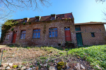 Old, abandoned, dilapidated house on hill against blue sky, weathered brick walls, broken gable roof with windows, metal and wooden doors, sunny day in Gulpen, South Limburg, Netherlands
