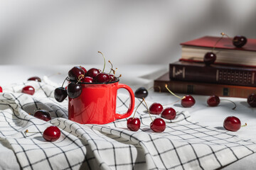 A red mug with ripe red cherries and a stack of books on a checkered napkin. Selective focus.