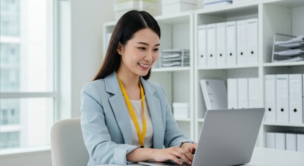 A smiling young woman in a light blue blazer sits at a desk in a bright office, using a laptop;  white shelves with files are in the background