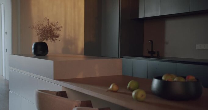 Modern kitchen interior with island, dining table, and bar stools. Warm sunlight illuminates fresh fruit in bowls. Plants add a touch of nature. Stylish and cozy atmosphere.