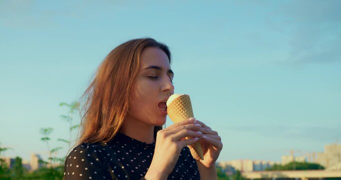 Stylish woman kissing ice cream cone at city overlook, woman cherishes vanilla flavor in golden hour light, scene continues with blurred skyline below and cream drip shimmering on cone edge.