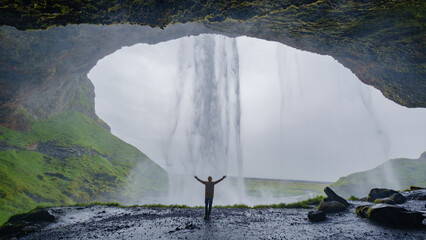 Seljalandsfoss waterfall tumbles from a jagged rock overhang, creating a misty curtain. A traveler stands beneath the waterfall in awe, surrounded by lush greenery and rugged terrain. © Fokke Baarssen